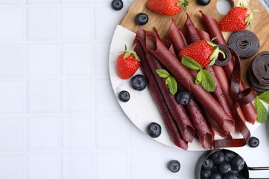 Photo of Tasty fruit leather rolls, strawberries, blueberries and mint on white tiled table, flat lay. Space for text