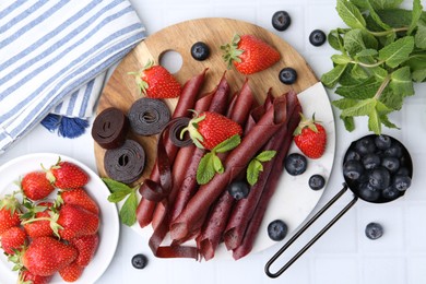 Photo of Tasty fruit leather rolls, strawberries, blueberries and mint on white tiled table, flat lay
