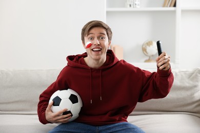 Photo of Emotional sports fan with remote control and soccer ball watching game indoors