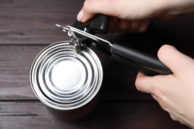 Photo of Woman opening tin with can opener at wooden table, top view