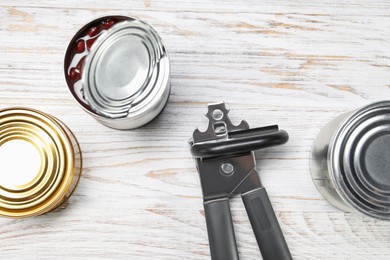 Photo of Can opener and tins on white wooden table, flat lay