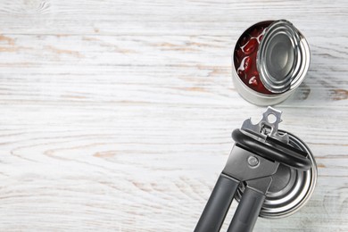 Photo of Can opener and tins on white wooden table, flat lay. Space for text