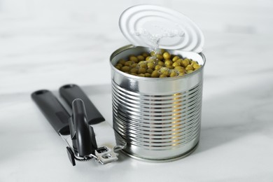 Photo of Can opener and tin with peas on white marble table, closeup