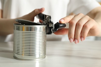Photo of Woman opening tin with can opener at white table, closeup