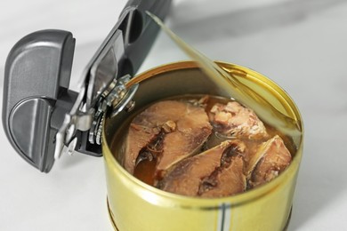 Photo of Can opener and tin with fish on white table, closeup
