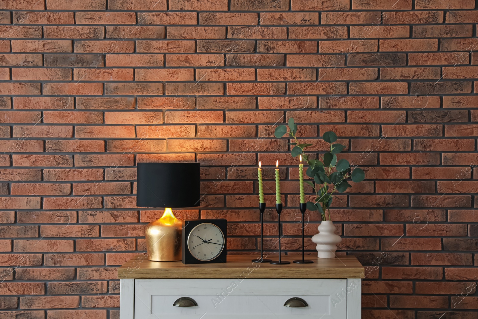 Candlesticks with burning candles on table near brick wall Photo of Candlesticks with burning candles on table near brick wall