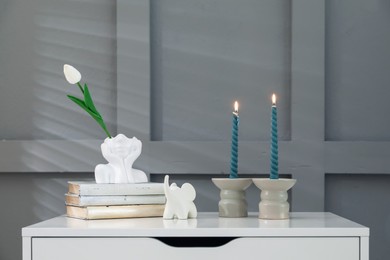 Photo of Candlesticks with burning candles on chest of drawers near grey wall