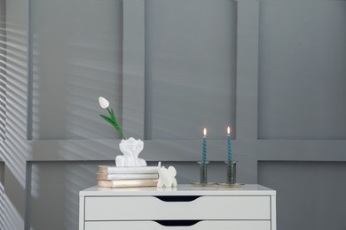 Photo of Candlesticks with burning candles on chest of drawers near grey wall