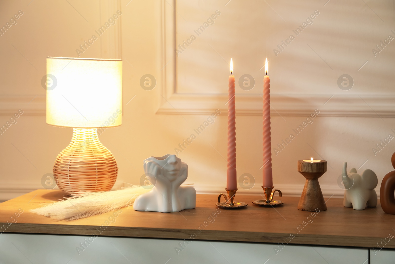 Candlesticks with burning candles on chest of drawers near white wall Photo of Candlesticks with burning candles on chest of drawers near white wall