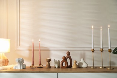 Photo of Candlesticks with burning candles on chest of drawers near white wall