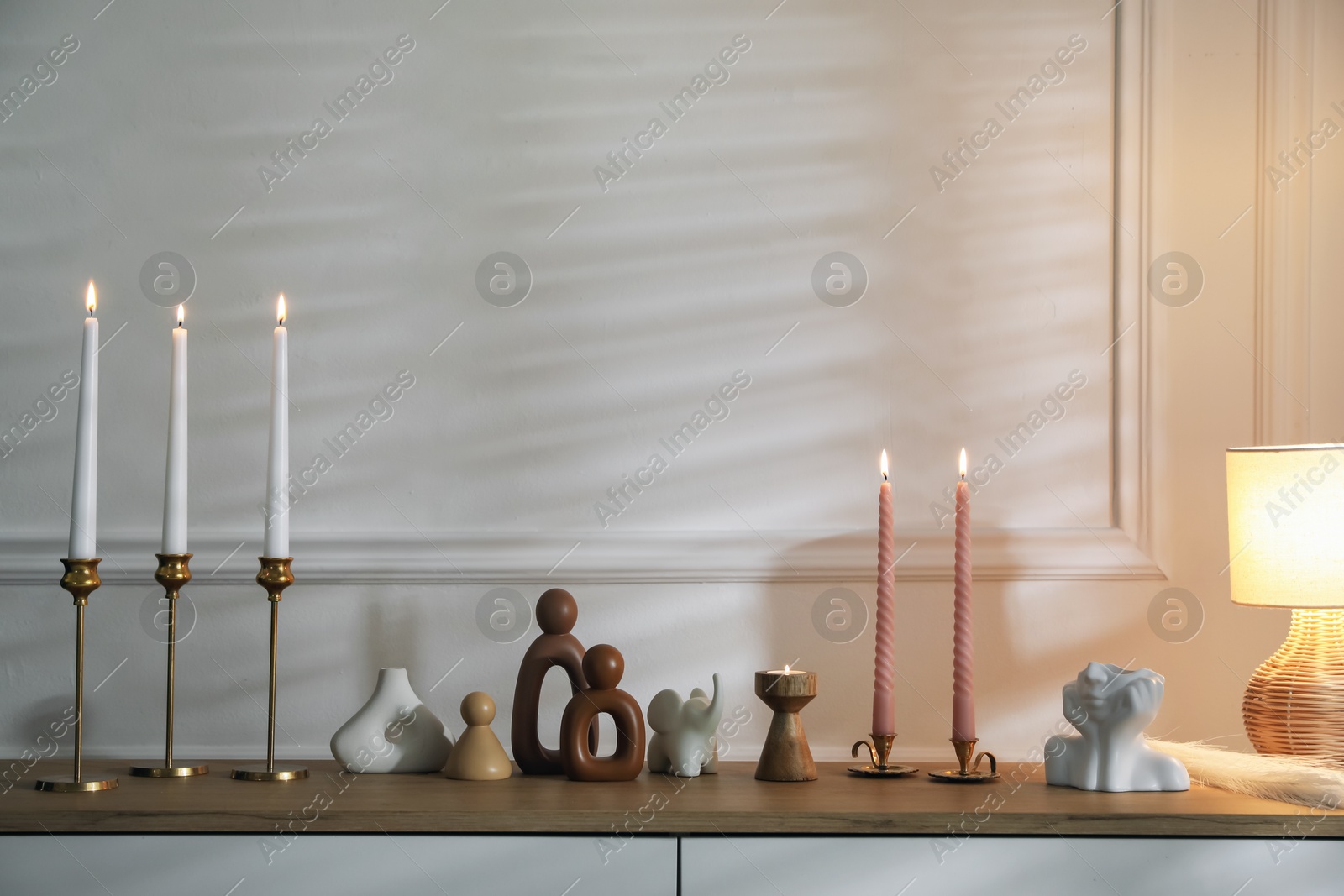 Candlesticks with burning candles on chest of drawers near white wall Photo of Candlesticks with burning candles on chest of drawers near white wall