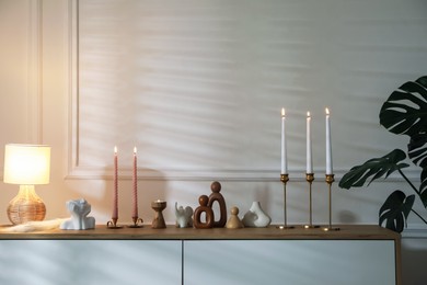 Photo of Candlesticks with burning candles on chest of drawers near white wall