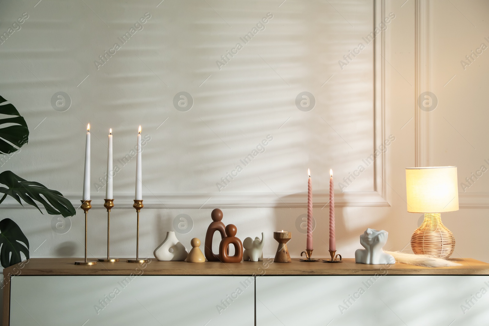 Candlesticks with burning candles on chest of drawers near white wall Photo of Candlesticks with burning candles on chest of drawers near white wall