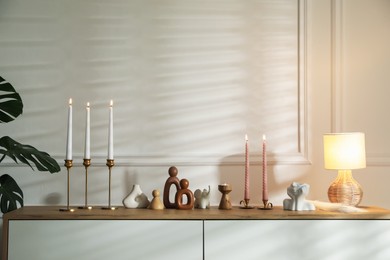 Photo of Candlesticks with burning candles on chest of drawers near white wall