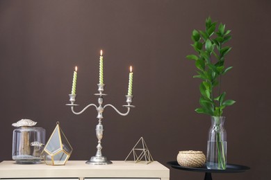 Photo of Elegant candlestick with burning candles and decor on cabinet near brown wall