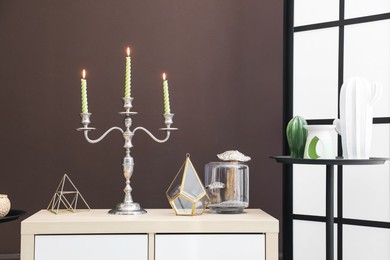 Photo of Elegant candlestick with burning candles and decor on cabinet near brown wall
