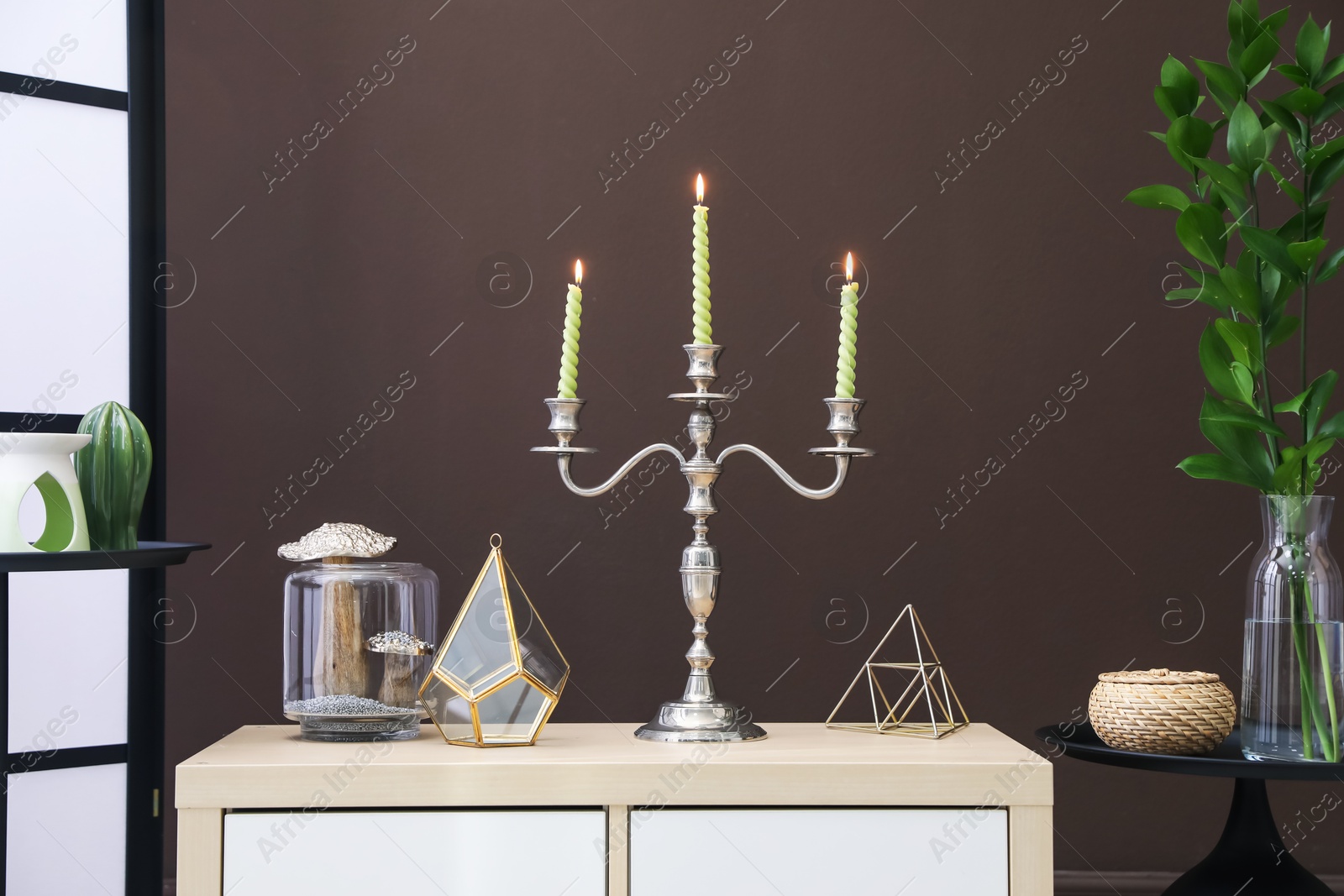 Elegant candlestick with burning candles and decor on cabinet near brown wall Photo of Elegant candlestick with burning candles and decor on cabinet near brown wall