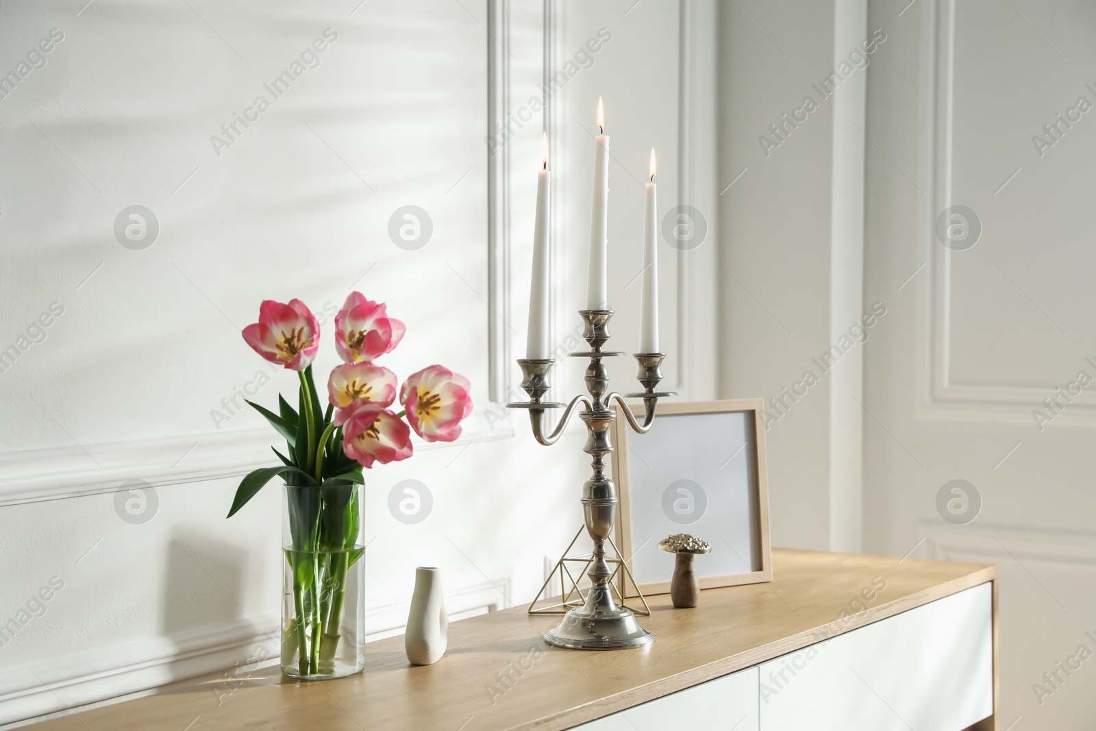 Elegant candlestick with burning candles and decor on chest of drawers near white wall Photo of Elegant candlestick with burning candles and decor on chest of drawers near white wall