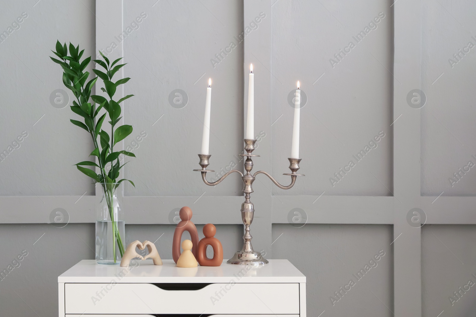 Elegant candlestick with burning candles and decor on chest of drawers near light wall Photo of Elegant candlestick with burning candles and decor on chest of drawers near light wall