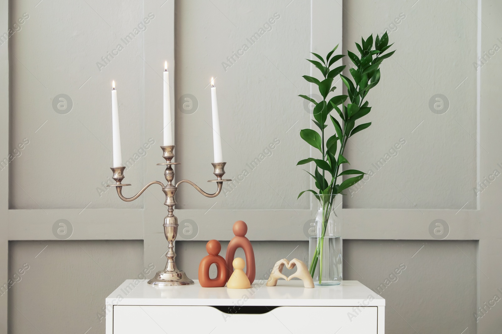 Elegant candlestick with burning candles and decor on chest of drawers near light wall Photo of Elegant candlestick with burning candles and decor on chest of drawers near light wall