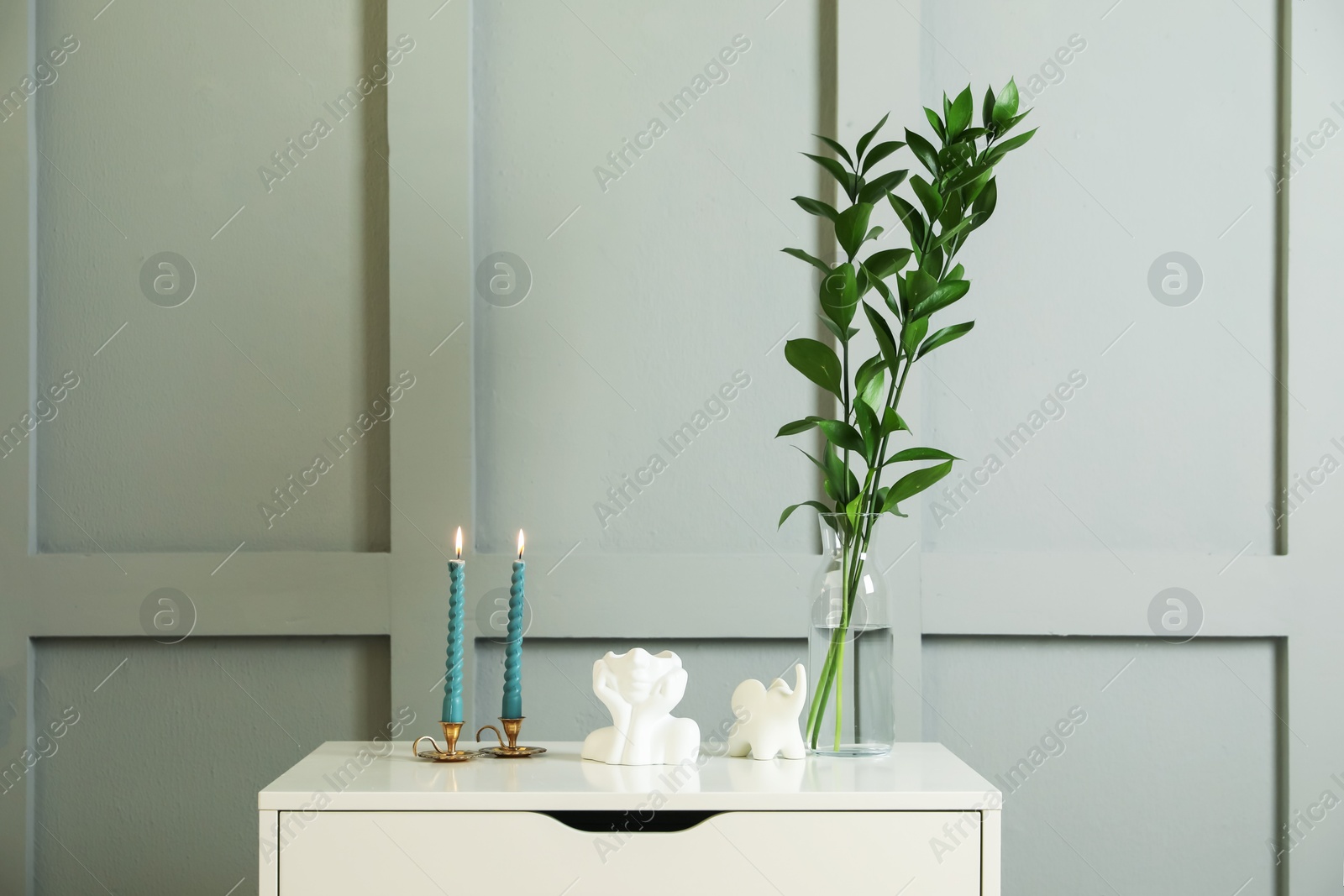 Candlesticks with burning candles on chest of drawers near light wall Photo of Candlesticks with burning candles on chest of drawers near light wall