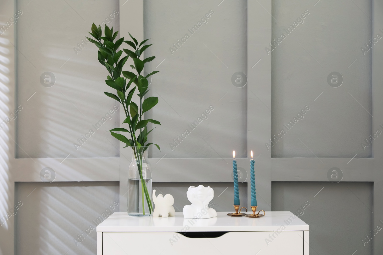 Candlesticks with burning candles on chest of drawers near light wall Photo of Candlesticks with burning candles on chest of drawers near light wall
