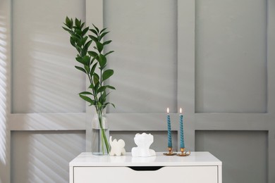Photo of Candlesticks with burning candles on chest of drawers near light wall