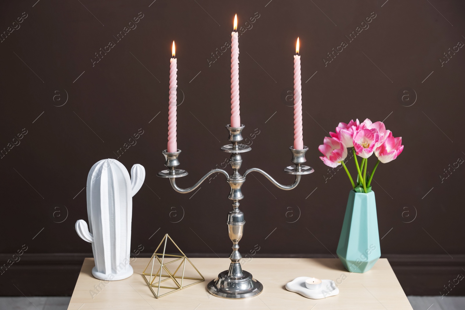 Elegant candlestick with burning candles and decor on table near brown wall Photo of Elegant candlestick with burning candles and decor on table near brown wall