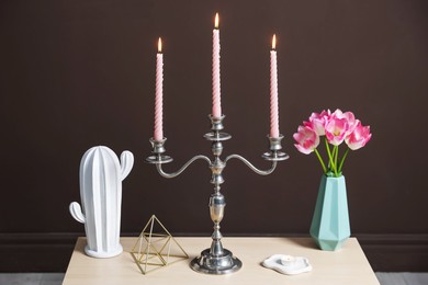 Photo of Elegant candlestick with burning candles and decor on table near brown wall