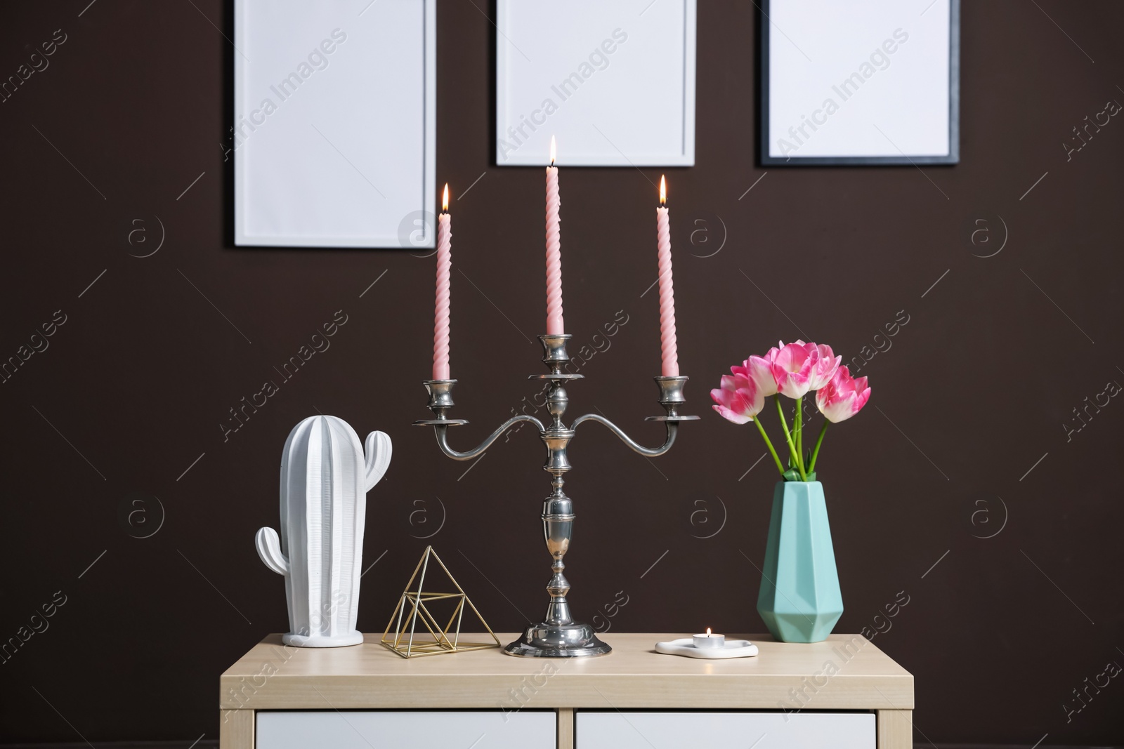 Elegant candlestick with burning candles and decor on cabinet near brown wall Photo of Elegant candlestick with burning candles and decor on cabinet near brown wall