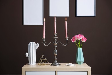 Photo of Elegant candlestick with burning candles and decor on cabinet near brown wall