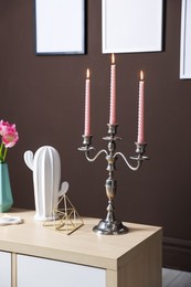 Photo of Elegant candlestick with burning candles and decor on cabinet near brown wall