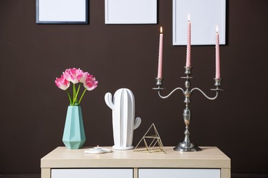 Photo of Elegant candlestick with burning candles and decor on cabinet near brown wall