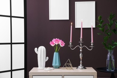 Photo of Elegant candlestick with burning candles and decor on cabinet near brown wall