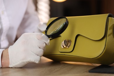 Photo of Appraiser with magnifying glass evaluating leather bag at table indoors, closeup