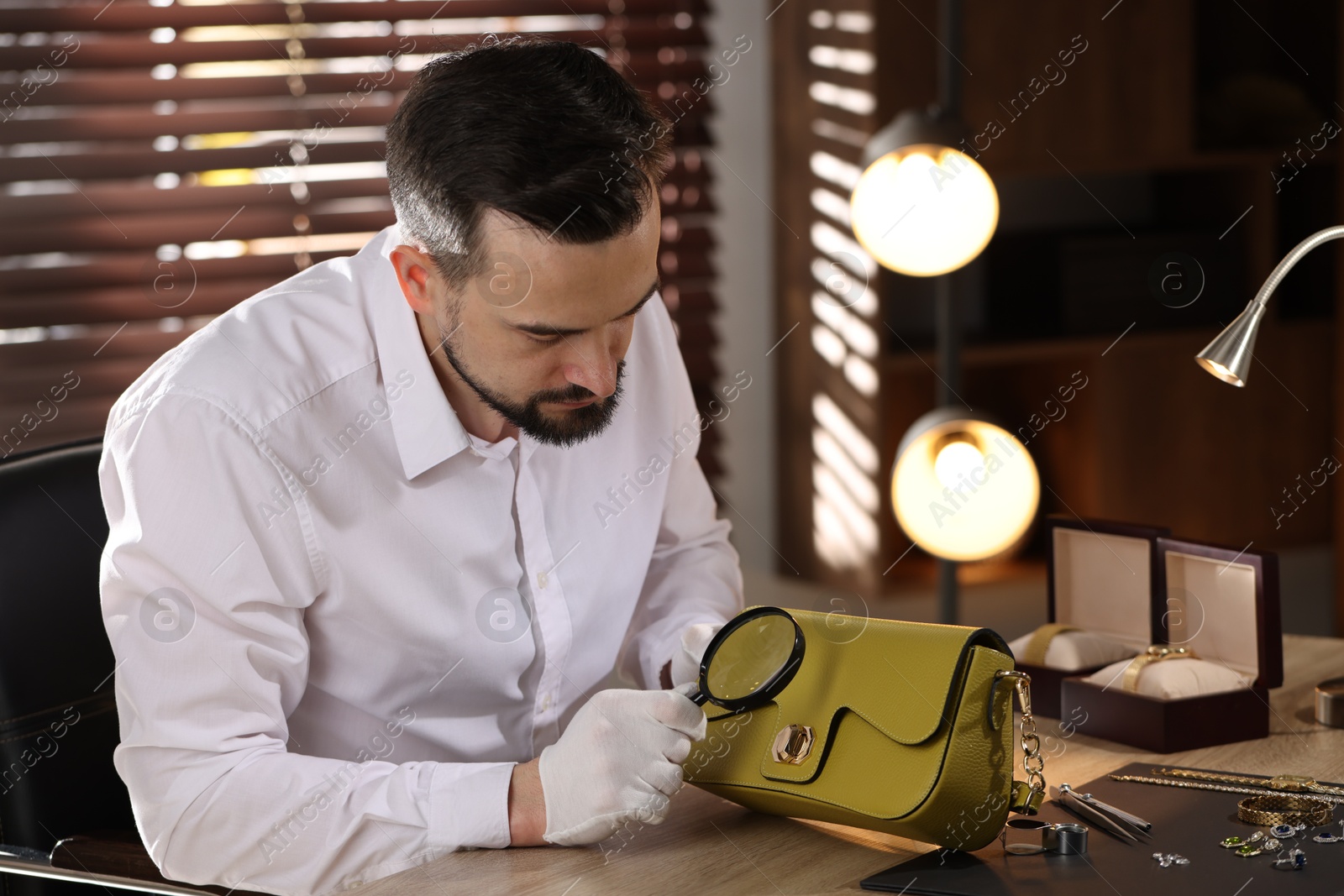 Appraiser with magnifying glass evaluating leather bag at table indoors Photo of Appraiser with magnifying glass evaluating leather bag at table indoors