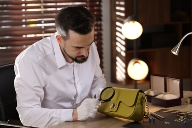 Photo of Appraiser with magnifying glass evaluating leather bag at table indoors