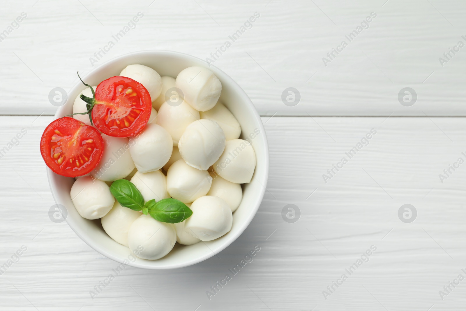 Tasty mozzarella cheese balls in bowl, basil and tomato on white wooden table, top view. Space for text Photo of Tasty mozzarella cheese balls in bowl, basil and tomato on white wooden table, top view. Space for text