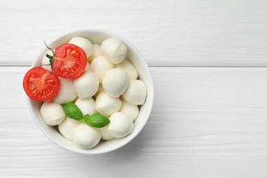 Photo of Tasty mozzarella cheese balls in bowl, basil and tomato on white wooden table, top view. Space for text