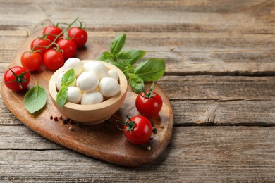 Photo of Tasty mozzarella cheese balls in bowl, basil, peppercorns and tomatoes on wooden table, space for text
