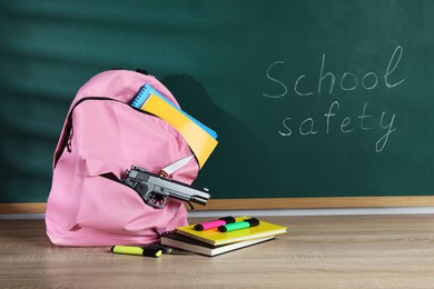 Photo of Backpack with gun, knife and stationery on wooden table near blackboard. School safety