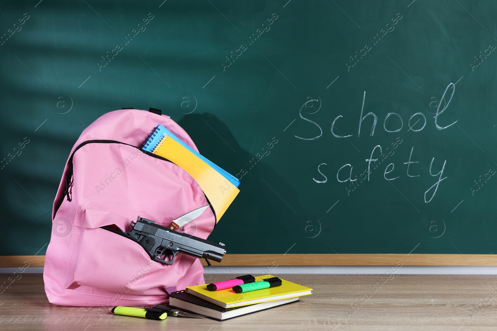 Backpack with gun, knife and stationery on wooden table near blackboard. School safety Photo of Backpack with gun, knife and stationery on wooden table near blackboard. School safety