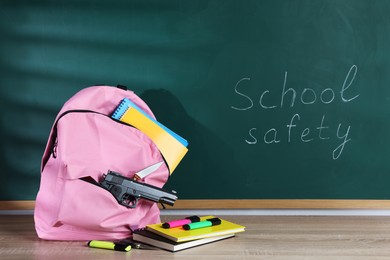 Photo of Backpack with gun, knife and stationery on wooden table near blackboard. School safety