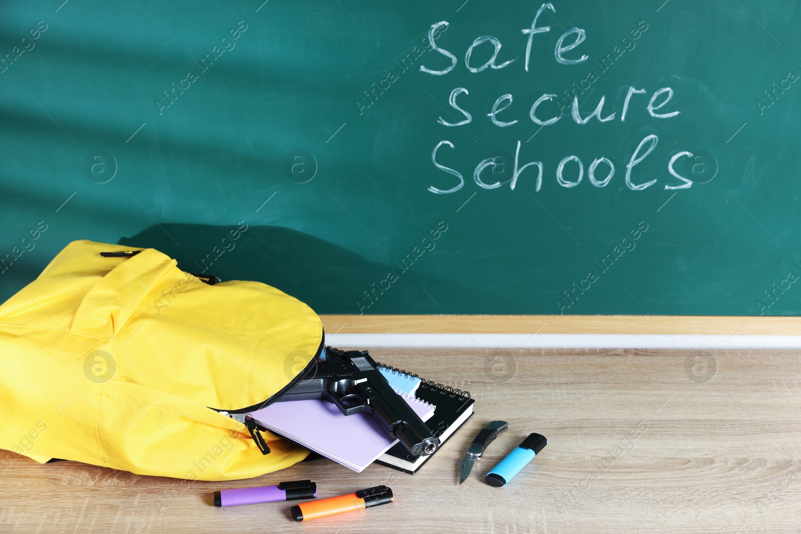 Backpack, gun, knife and stationery on wooden table near blackboard with words Safe Secure Schools Photo of Backpack, gun, knife and stationery on wooden table near blackboard with words Safe Secure Schools