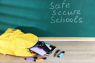 Photo of Backpack, gun, knife and stationery on wooden table near blackboard with words Safe Secure Schools