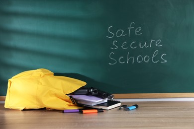 Photo of Backpack, gun, knife and stationery on wooden table near blackboard with words Safe Secure Schools