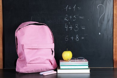 Photo of Blackboard with addition problems to solve, backpack, notebooks, highlighters, glasses and apple on black table