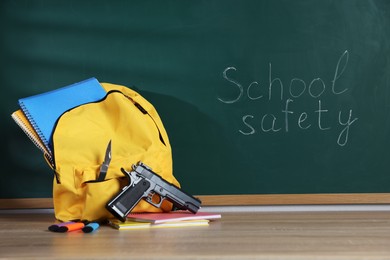 Photo of Backpack with gun, knife and stationery on wooden table near blackboard. School safety