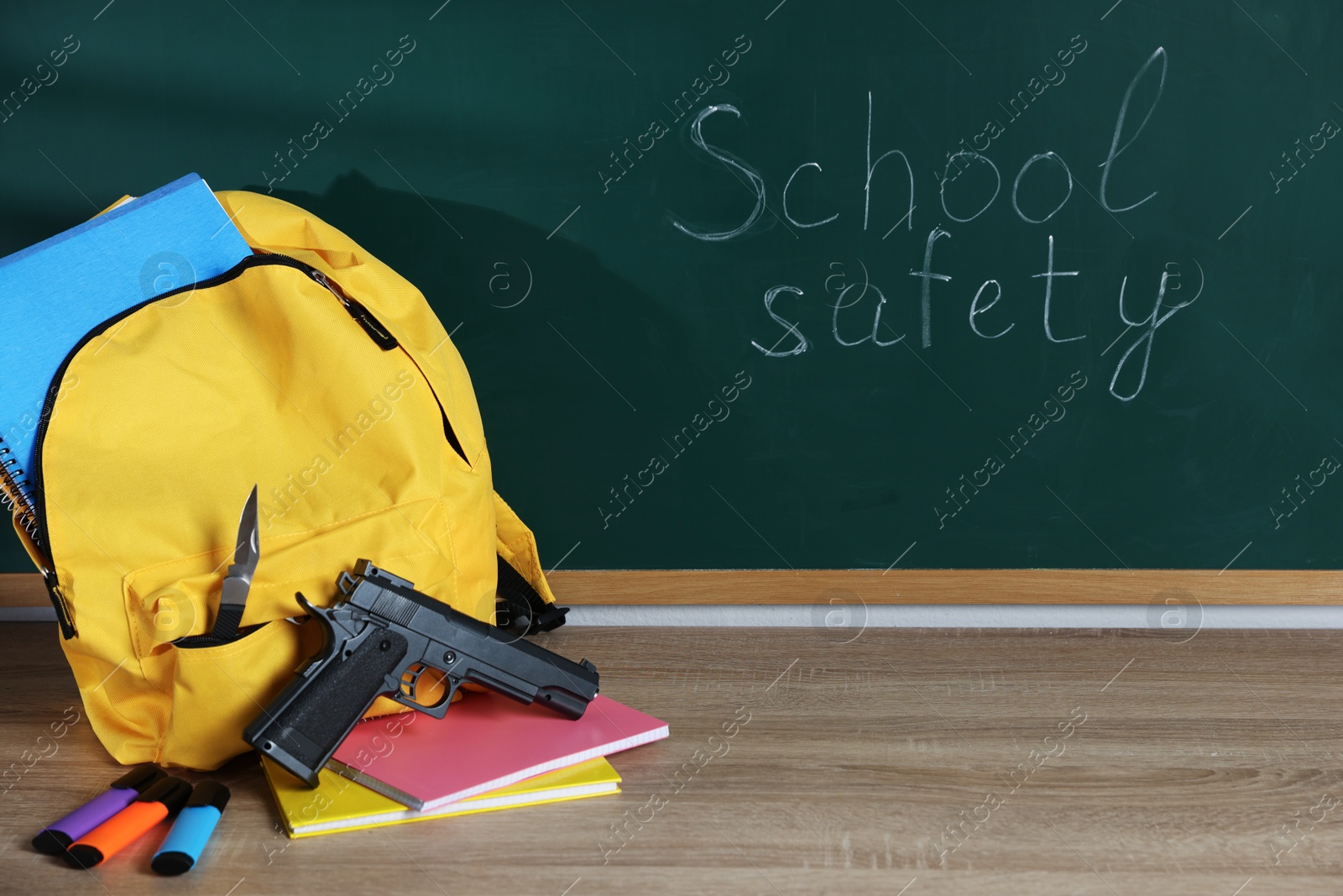 Backpack with gun, knife and stationery on wooden table near blackboard. School safety Photo of Backpack with gun, knife and stationery on wooden table near blackboard. School safety