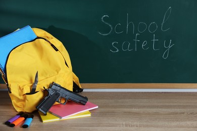 Photo of Backpack with gun, knife and stationery on wooden table near blackboard. School safety